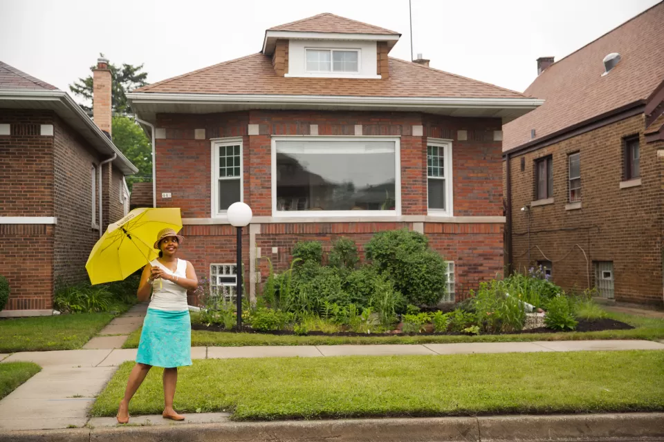 A Black woman stands in front of a home holding an umbrella and smiling.